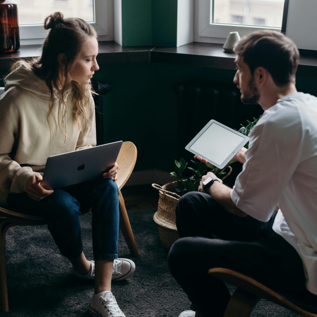 Two people sit facing each other indoors; one holds a laptop, the other holds a tablet. They appear to be engaged in conversation.