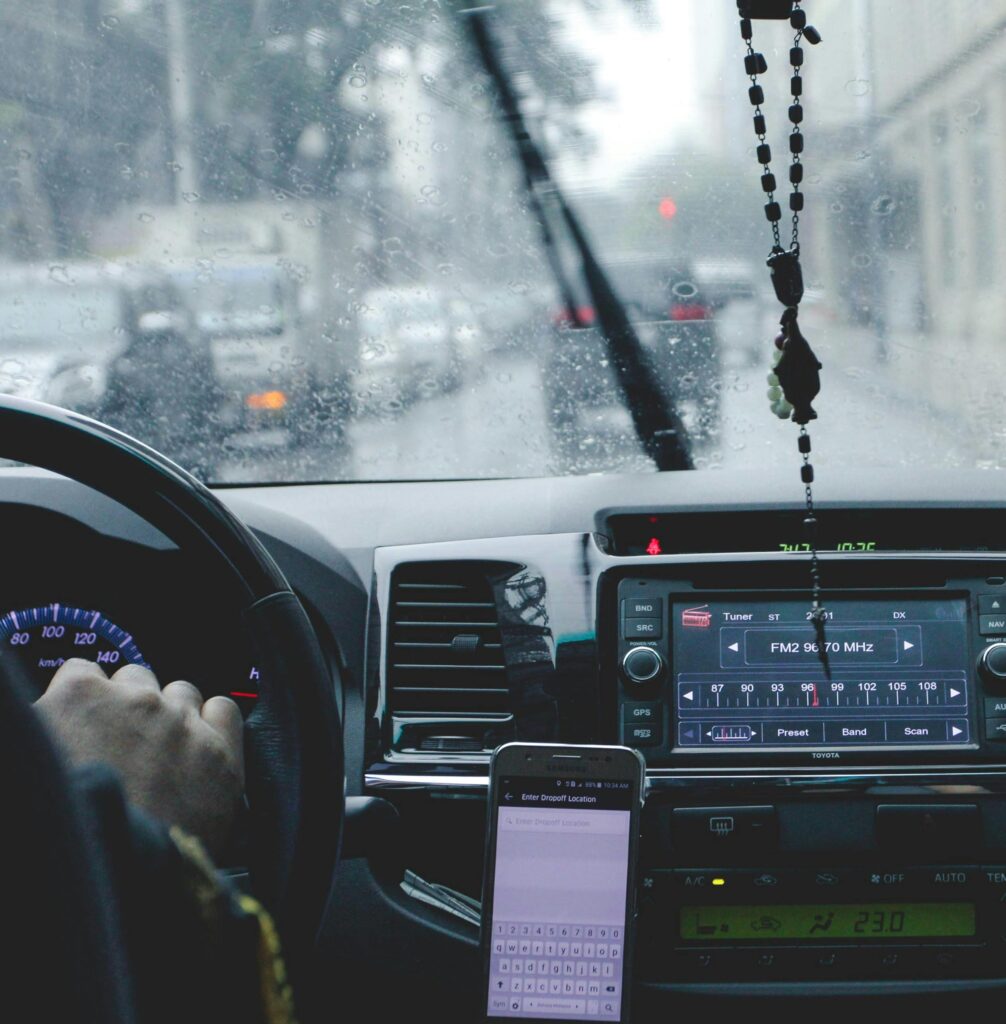 A person drives a car on a rainy day with a phone mounted near the dashboard and a rosary hanging from the rearview mirror. The radio display shows an FM station.