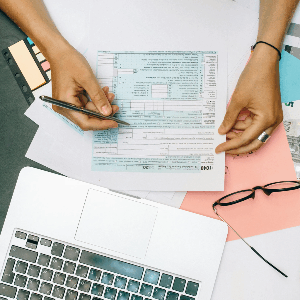 Person filling out a tax form with a pen, surrounded by papers, a laptop, sticky notes, and a pink folder on a desk.