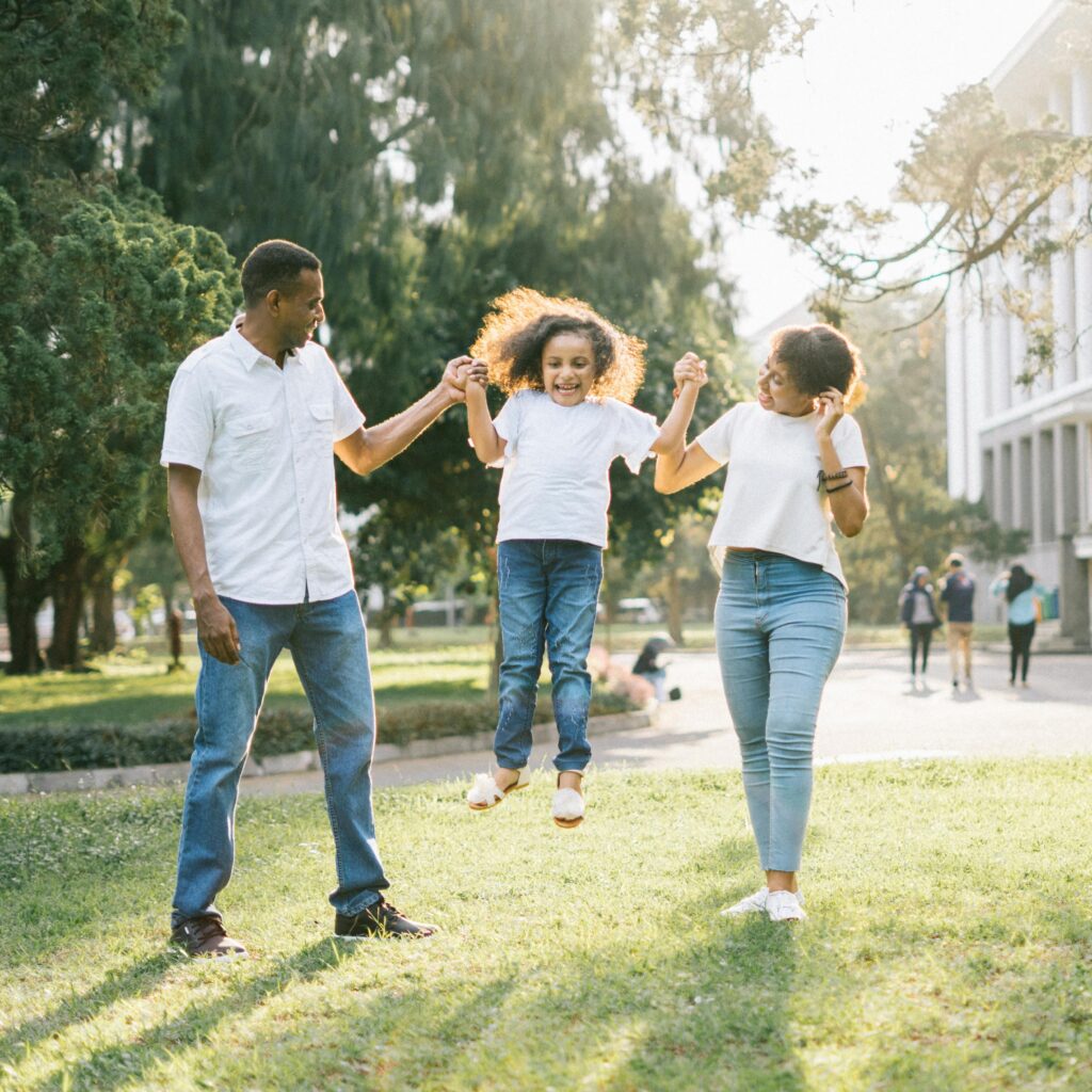 Two adults hold a child by the hands, lifting them off the ground, while standing on grass in a park with trees and people in the background.